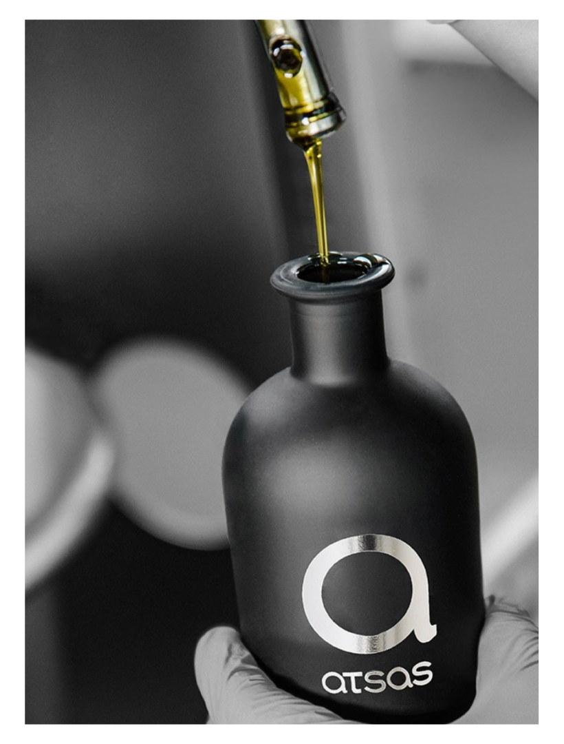 Black bottle with 'atsas' logo being filled with a olive oil using a dropper.